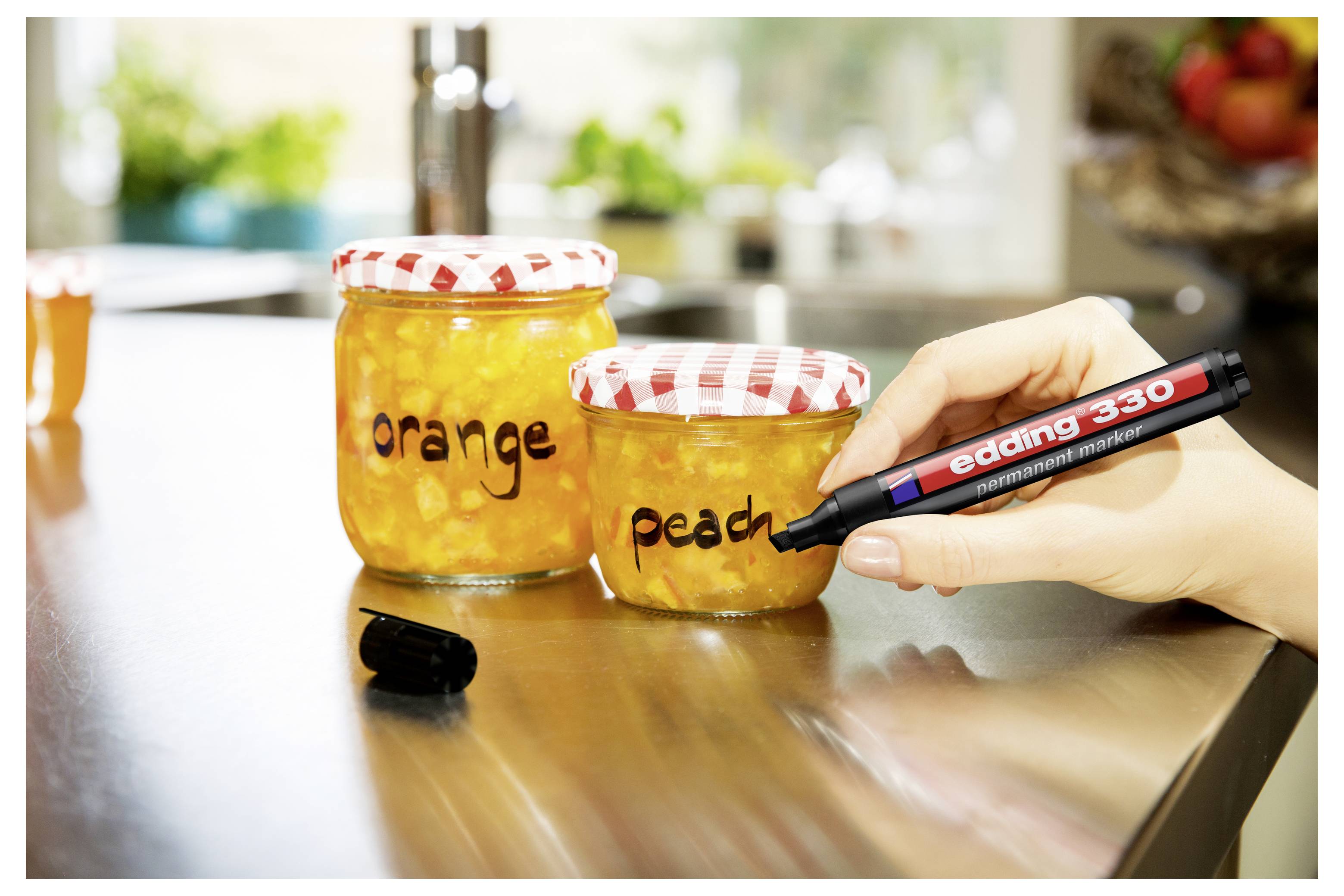 Eine Hand beschriftet ein Marmeladenglas mit "Pfirsich" mit einem schwarzen Marker neben einem anderen Glas mit der Aufschrift "Orange" auf einer Küchenarbeitsplatte.