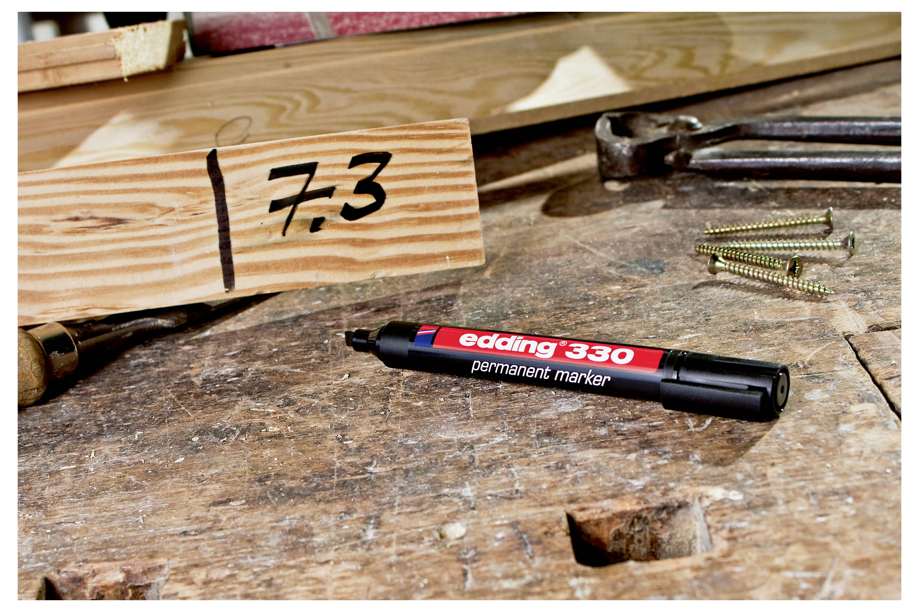 Permanentmarker auf einer Werkbank neben einem Holzblock mit der Aufschrift „7.3", mit Schrauben und Handwerkzeugen im Hintergrund.