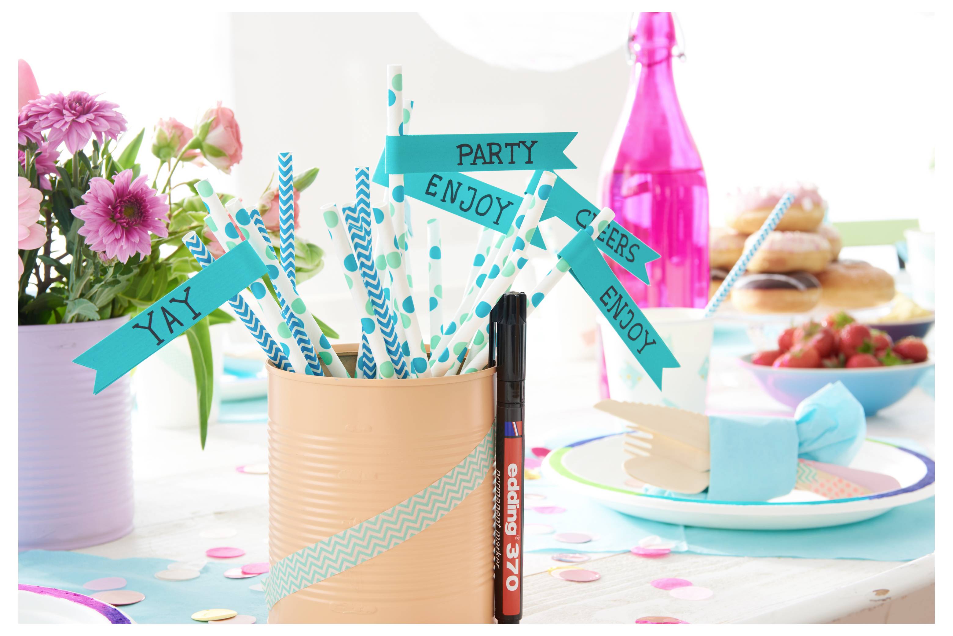 Partytischtischdekoration mit blaugrünen gestreiften Strohhalmen und Bannern mit den Aufschriften „YAY", „PARTY", „ENJOY" in einem pfirsichfarbenen Becher, sowie Desserts, Blumen und einer rosafarbenen Flasche.