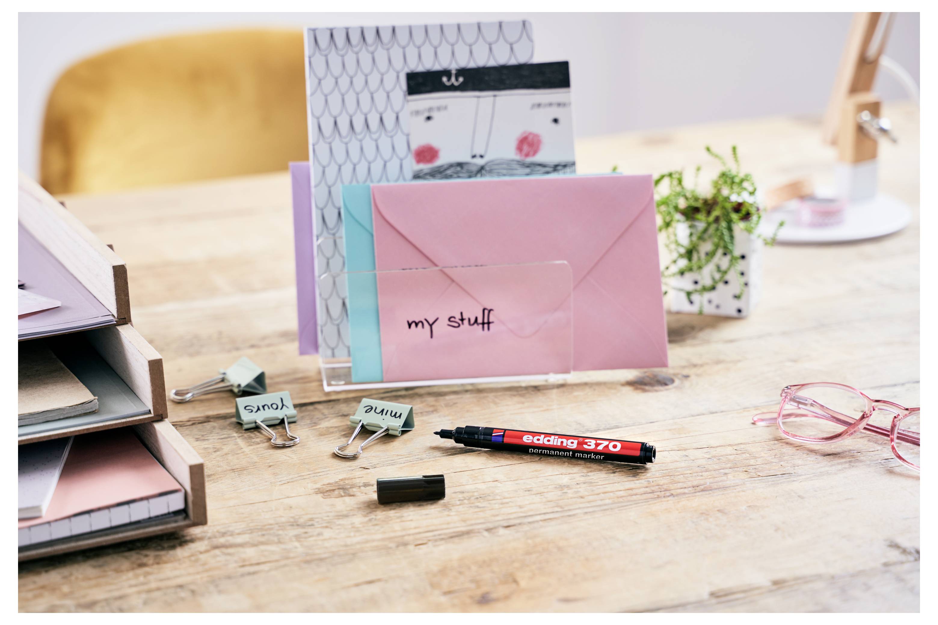 Schreibtisch mit rosa und türkisfarbenen Umschlägen, Büroklammern mit den Aufschriften „smy" und „alpha", einem roten Marker, rosa Brille und einem kleinen Pflanzenpflanzorganizer.