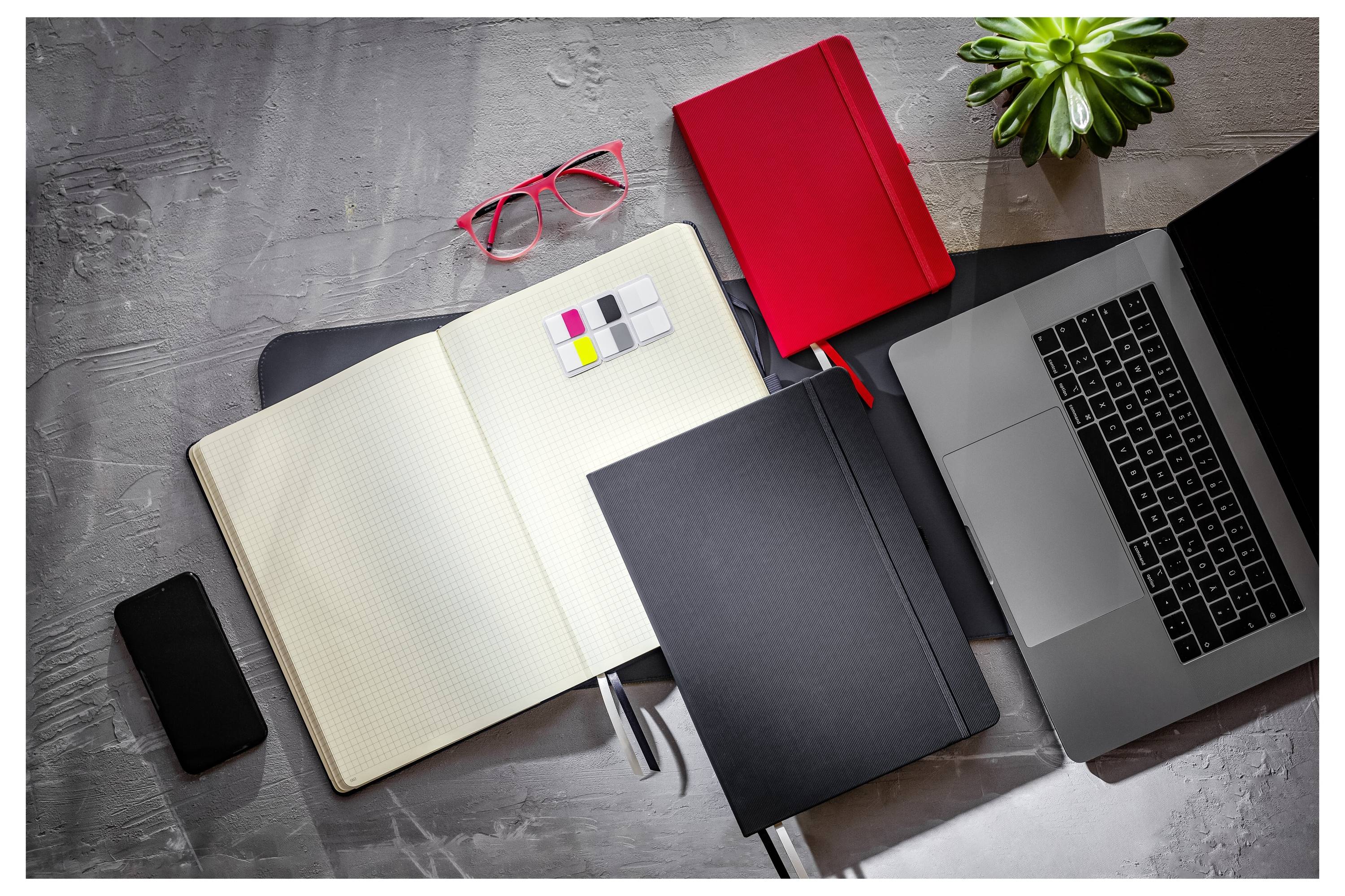 Draufsicht eines Arbeitsplatzes mit einem offenen Notizbuch, geschlossenem Ordner, Laptop, roter Brille, Telefon, rotem Notizbuch und einer kleinen Pflanze auf einem dunklen Schreibtisch.