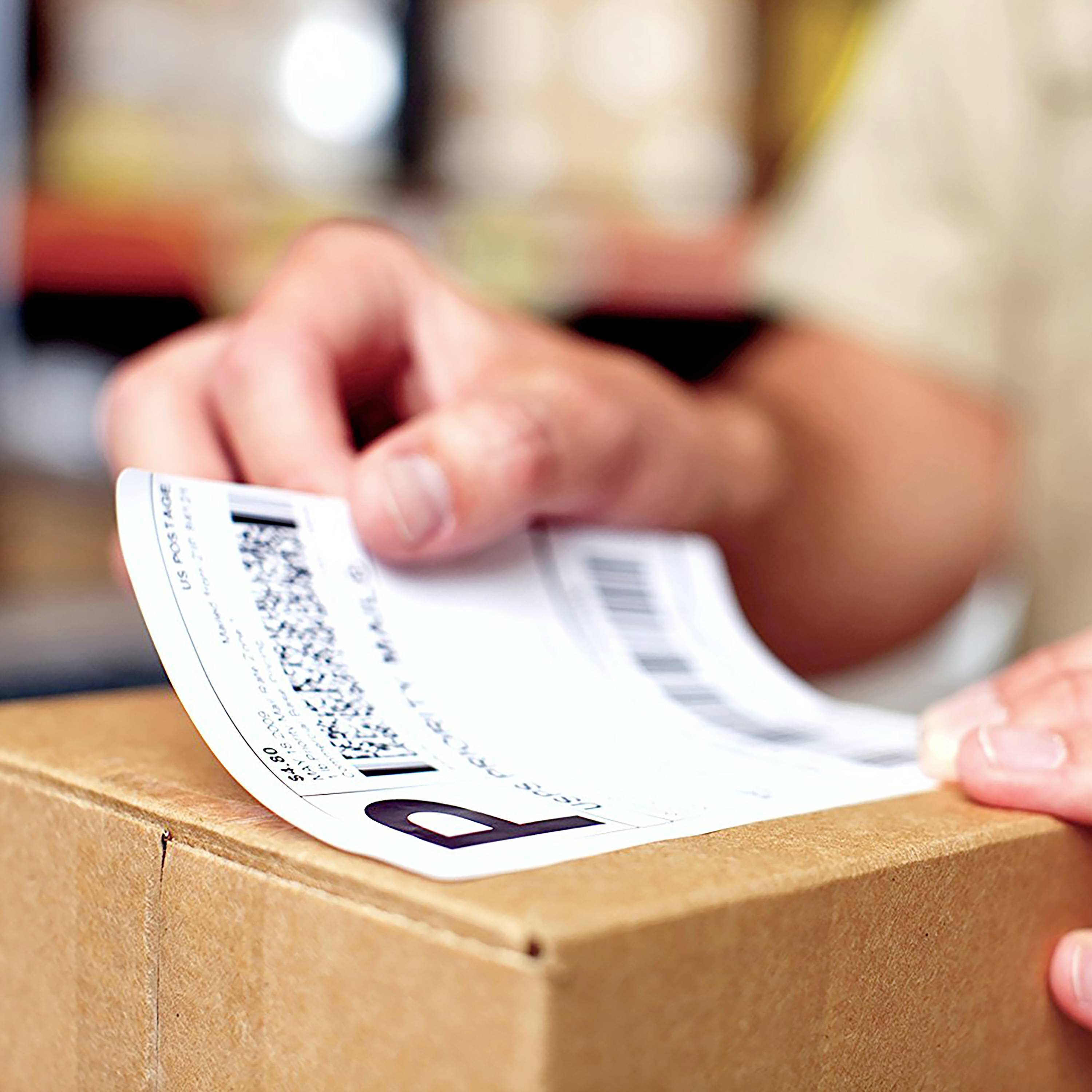 Eine Hand hält ein Versandetikett auf einem braunen Paket. Der Hintergrund ist unscharf.