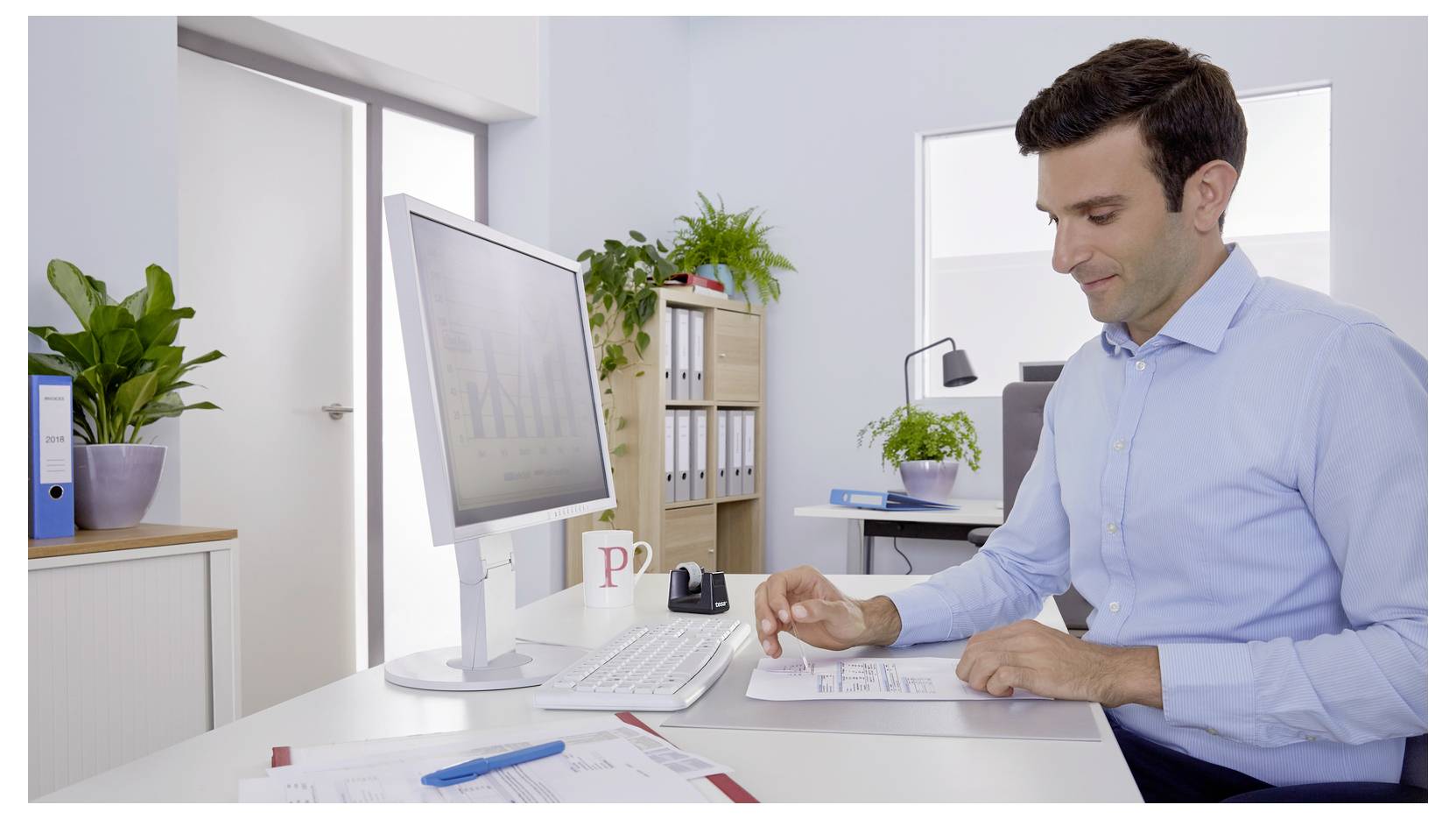 Ein Mann in einem blauen Hemd sitzt an einem Büroschreibtisch und überprüft ein Dokument. Ein Computermonitor, Papiere und Pflanzen sind im Raum sichtbar.