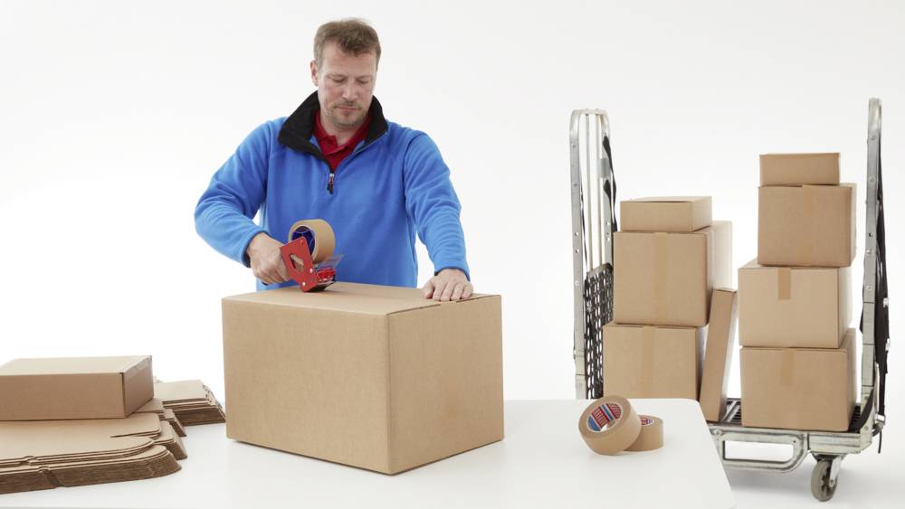 Ein Mann in blauem Pullover verklebt einen großen Karton mit Klebeband. Im Hintergrund stehen weitere Kartons auf einem Rollwagen.