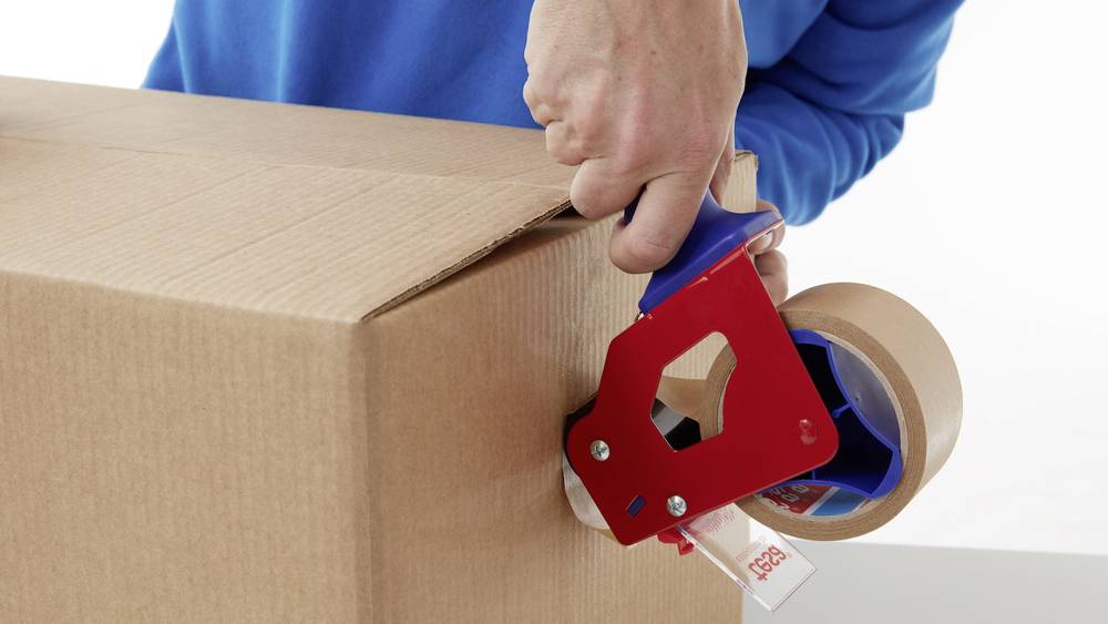 Eine Person verschließt einen Karton mit Klebeband und verwendet dafür einen roten Abroller.