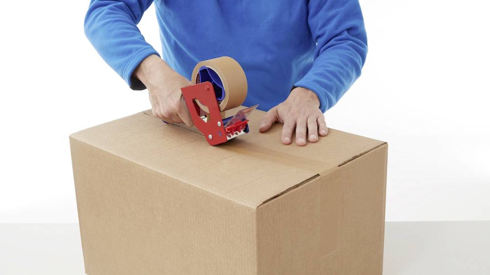 Person verpackt einen Karton mit Klebeband. Die Person trägt einen blauen Pullover und nutzt einen roten Handabroller.
