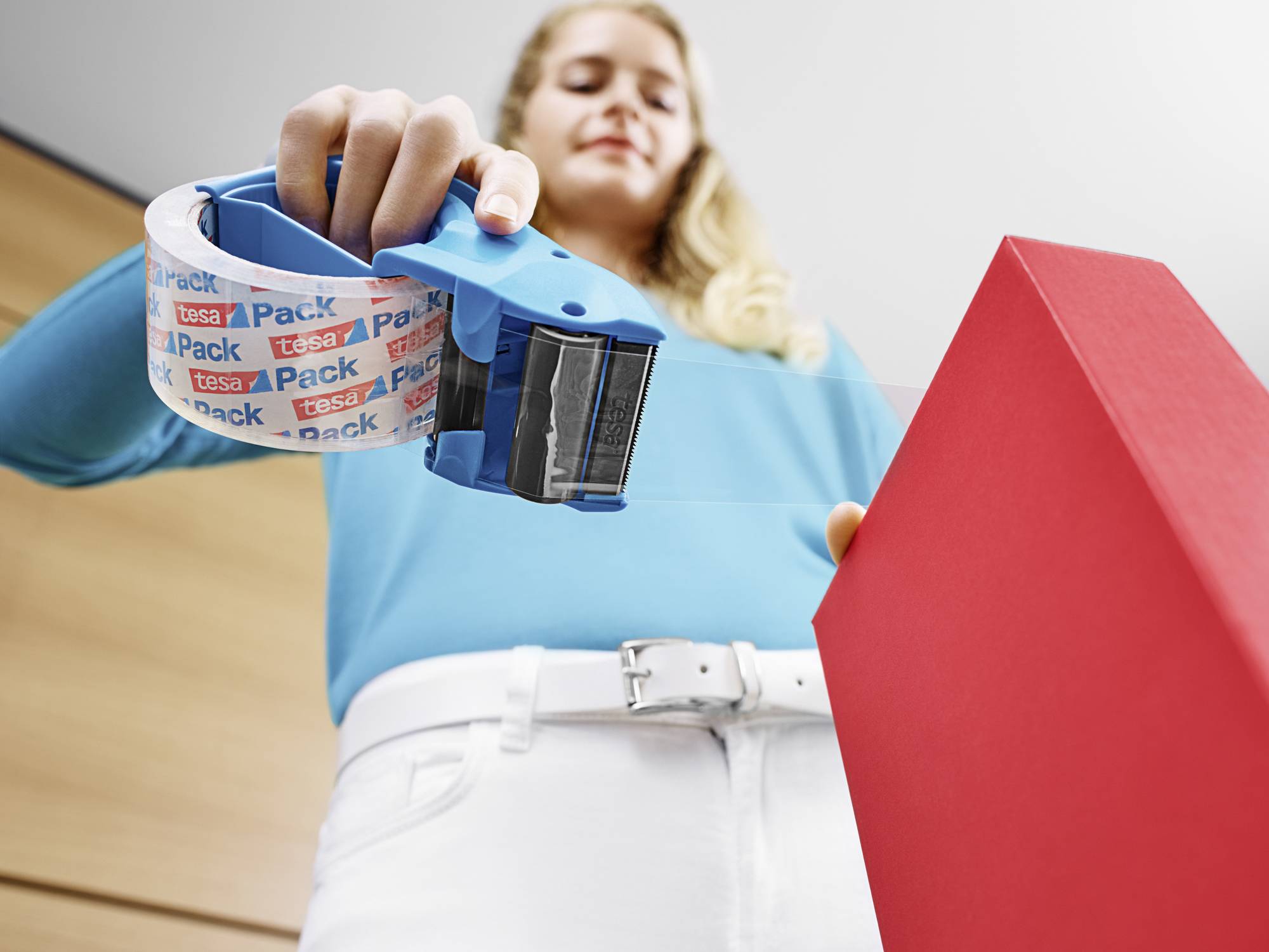 Eine Frau im blauen Pullover versiegelt ein rotes Paket mit einem blauen Klebebandspender. Das Klebeband ist bedruckt mit 'tesa Pack'.