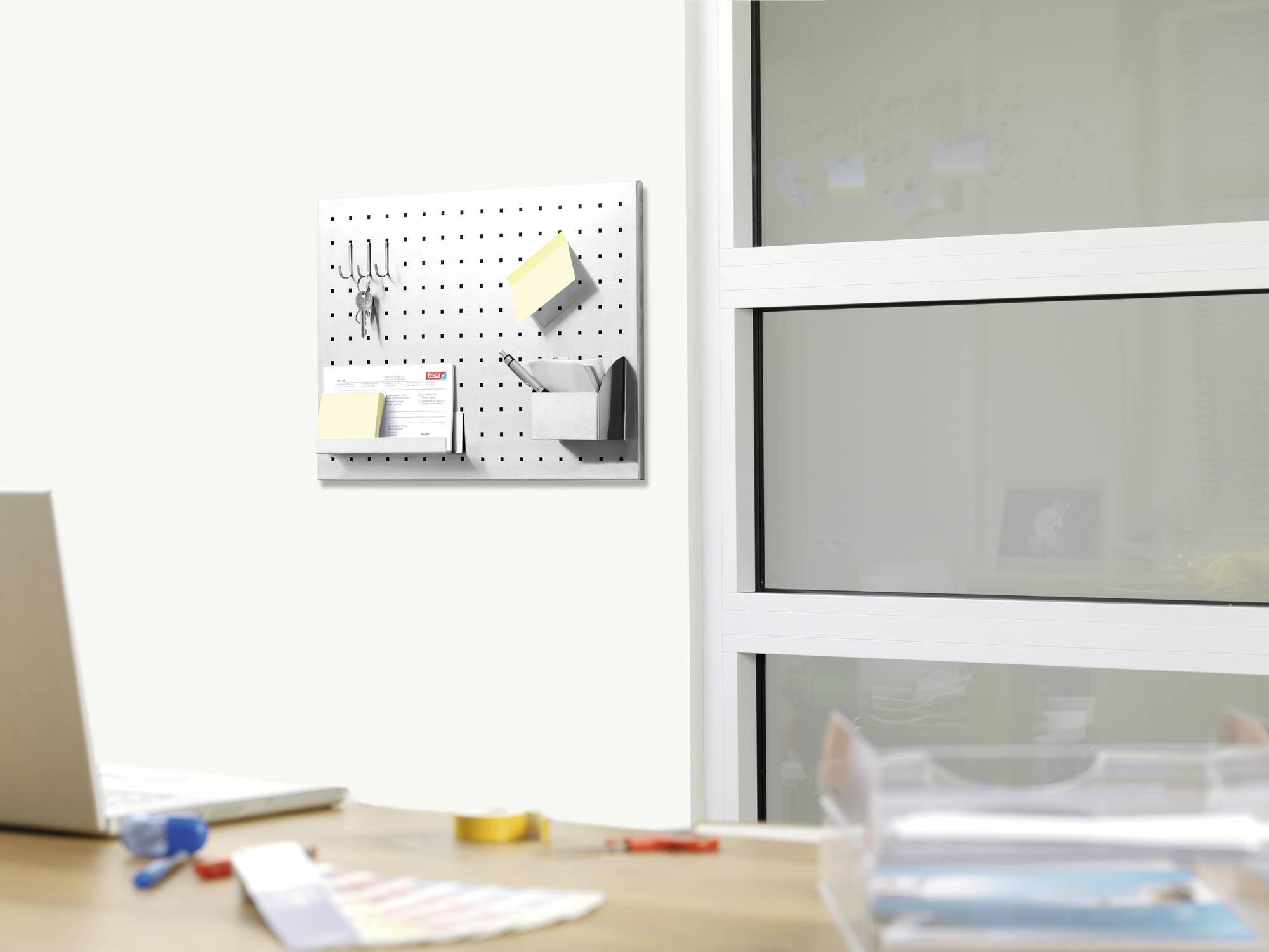Eine Schreibtischszene mit einem Laptop im Vordergrund und einem perforierten Wand-Organizer mit Büroartikeln an der Wand im Hintergrund.