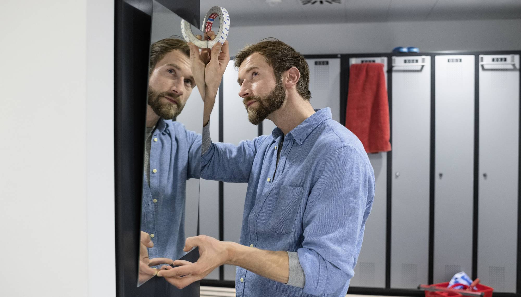 Ein Mann in einem blauen Hemd betrachtet sich im Spiegel in einem Umkleideraum. Im Hintergrund sind Schließfächer und ein rotes Handtuch.