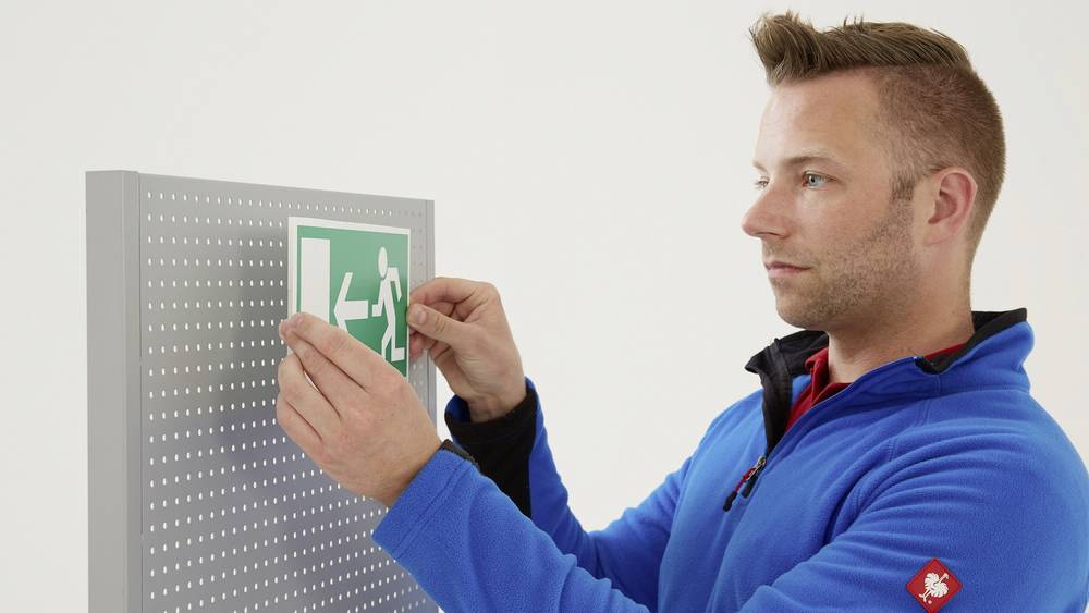 Ein Mann in blauer Jacke befestigt ein grünes Notausgangsschild an einer perforierten Tafel.