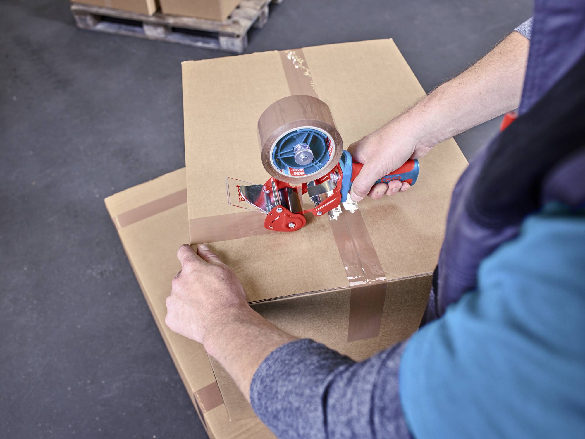 Eine Person verschließt einen Karton mit Packband. Der Karton steht auf einem weiteren Karton auf einem Betonboden.