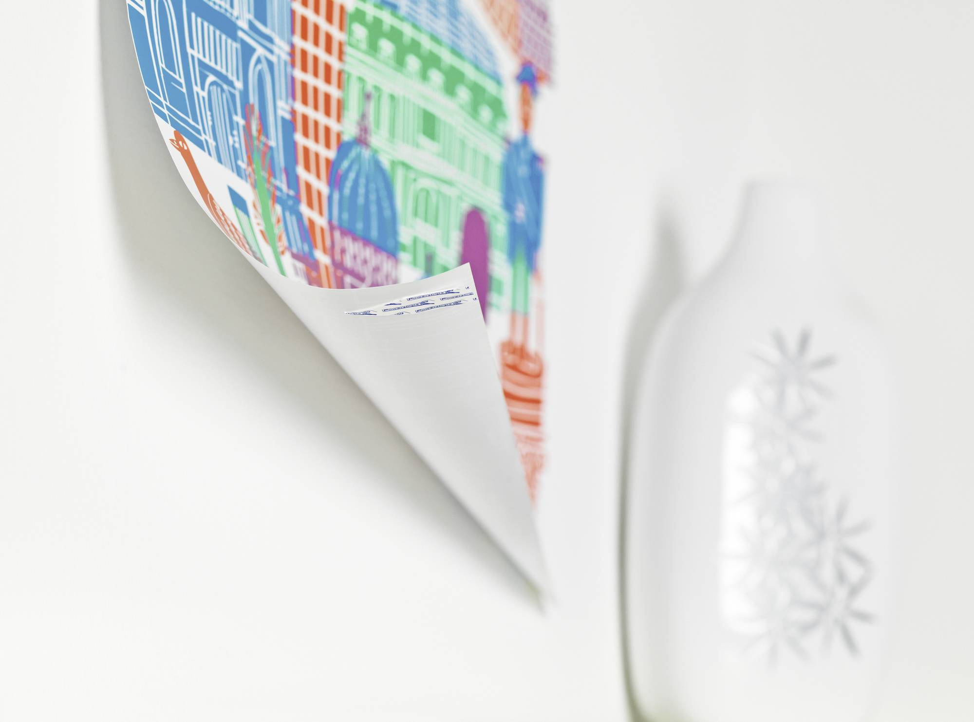 Farbiges Stadtmotiv-Poster hängt neben einer weißen Vase mit floralen Motiven auf einem weißen Hintergrund.