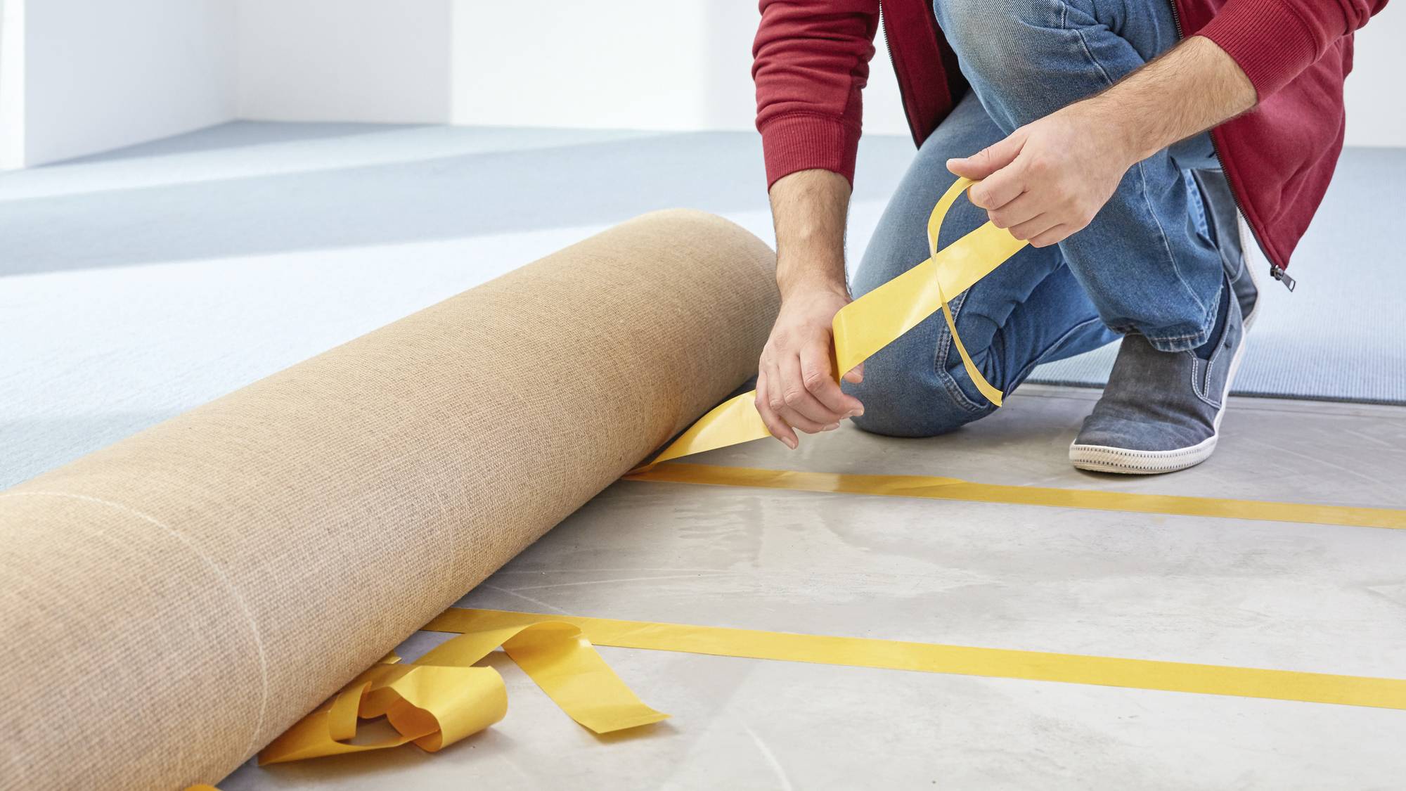 Person verlegt Teppichboden und verwendet gelbes Klebeband zum Fixieren. Nahaufnahme von Händen im Arbeitsprozess.