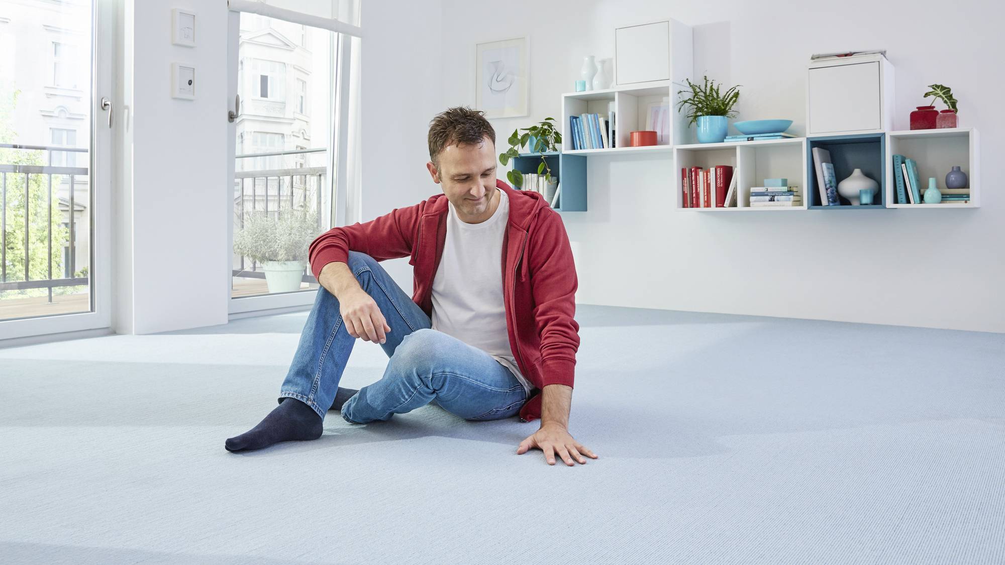 Ein Mann sitzt lächelnd auf einem hellblauen Teppichboden in einem modern eingerichteten Wohnzimmer mit Regalen im Hintergrund.