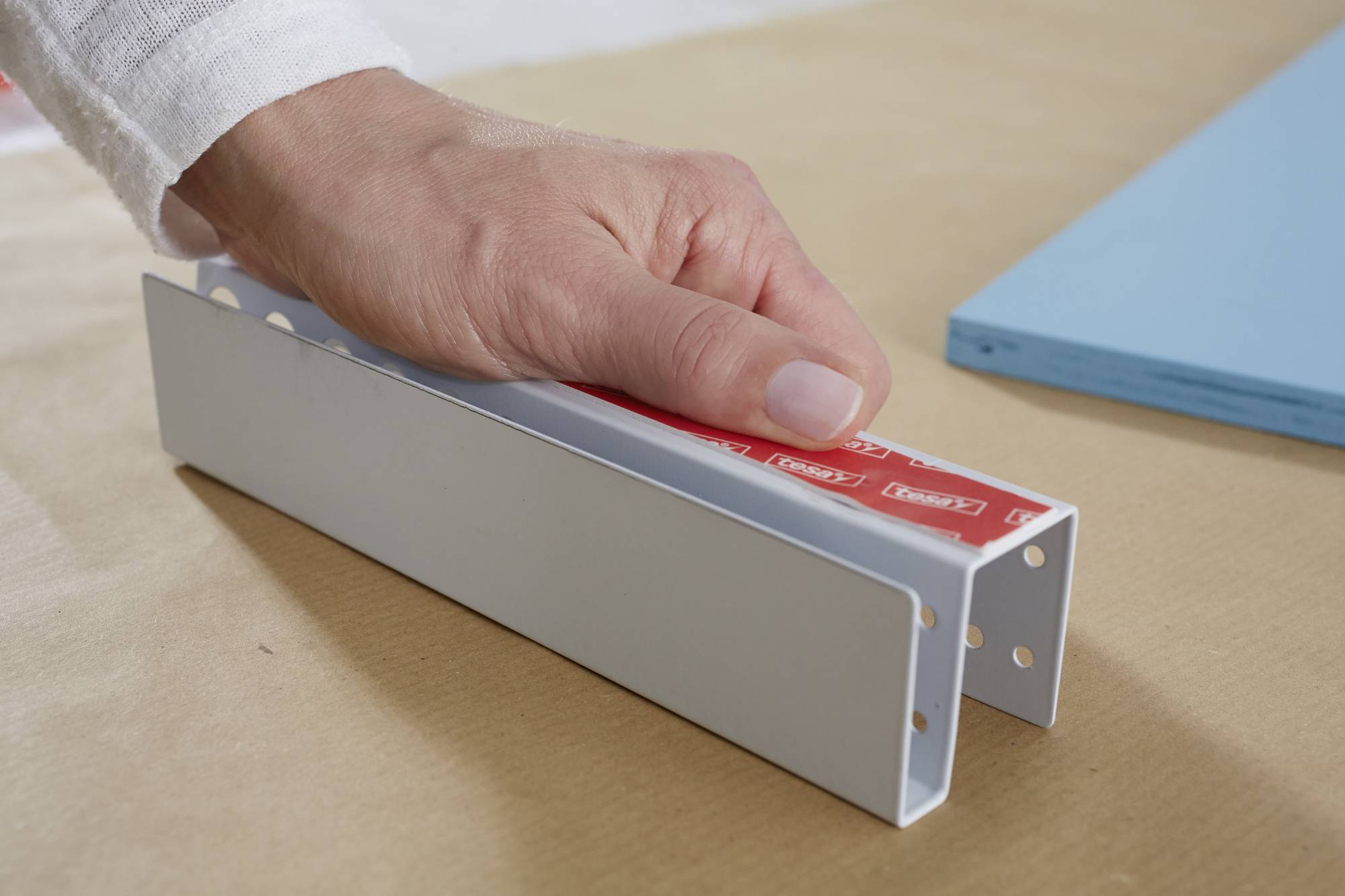 Eine Hand hält ein U-förmiges Metallstück mit rotem Klebeband, das auf einem Tisch liegt. Daneben liegt ein blaues Rechteck.