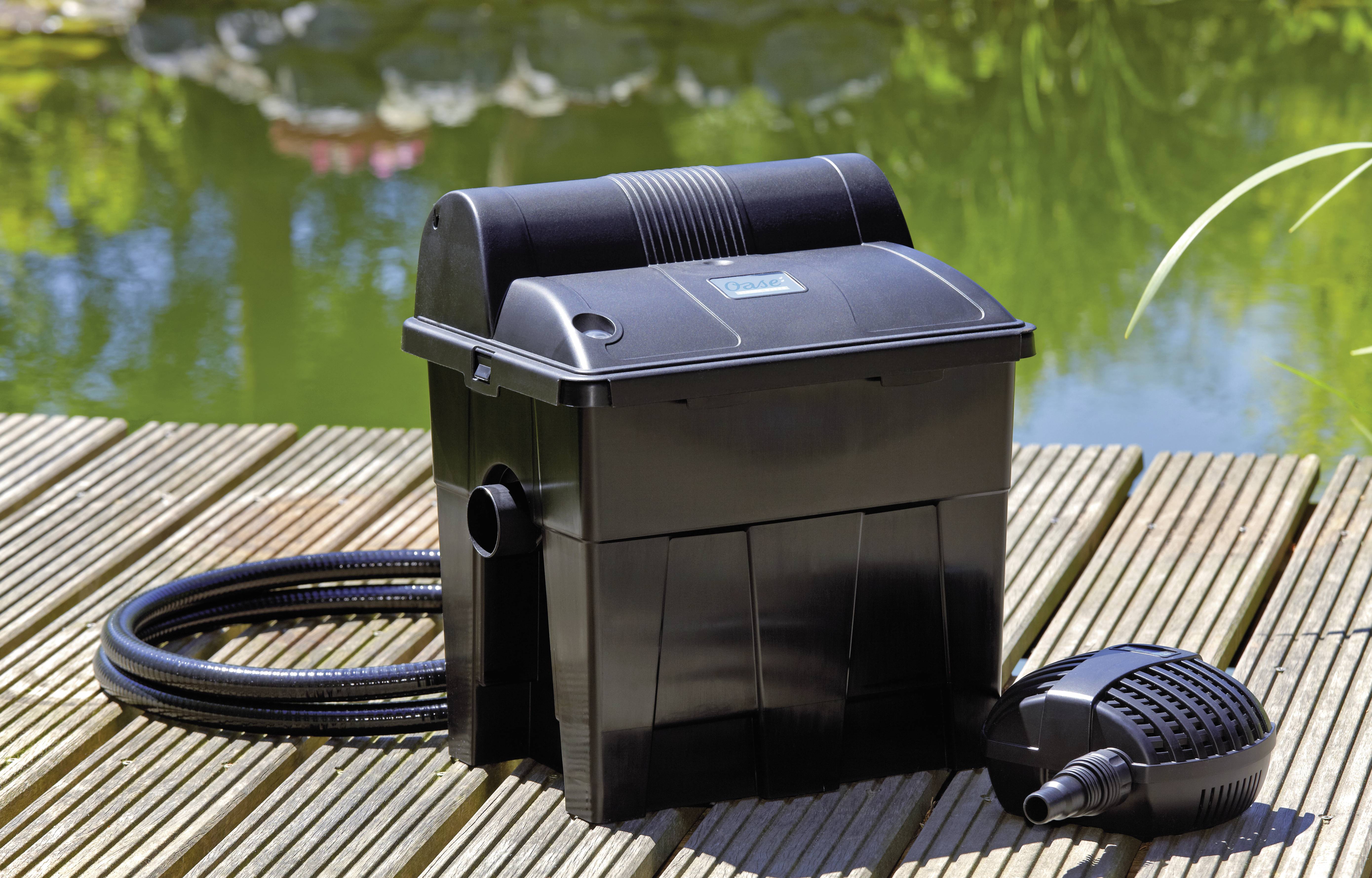 Ein schwarzer Teichfilter mit einem Schlauch und einer Pumpe auf einer Holzterrasse. Ein Teich mit Grünpflanzen ist im Hintergrund zu sehen und deutet auf eine Gartenumgebung hin.