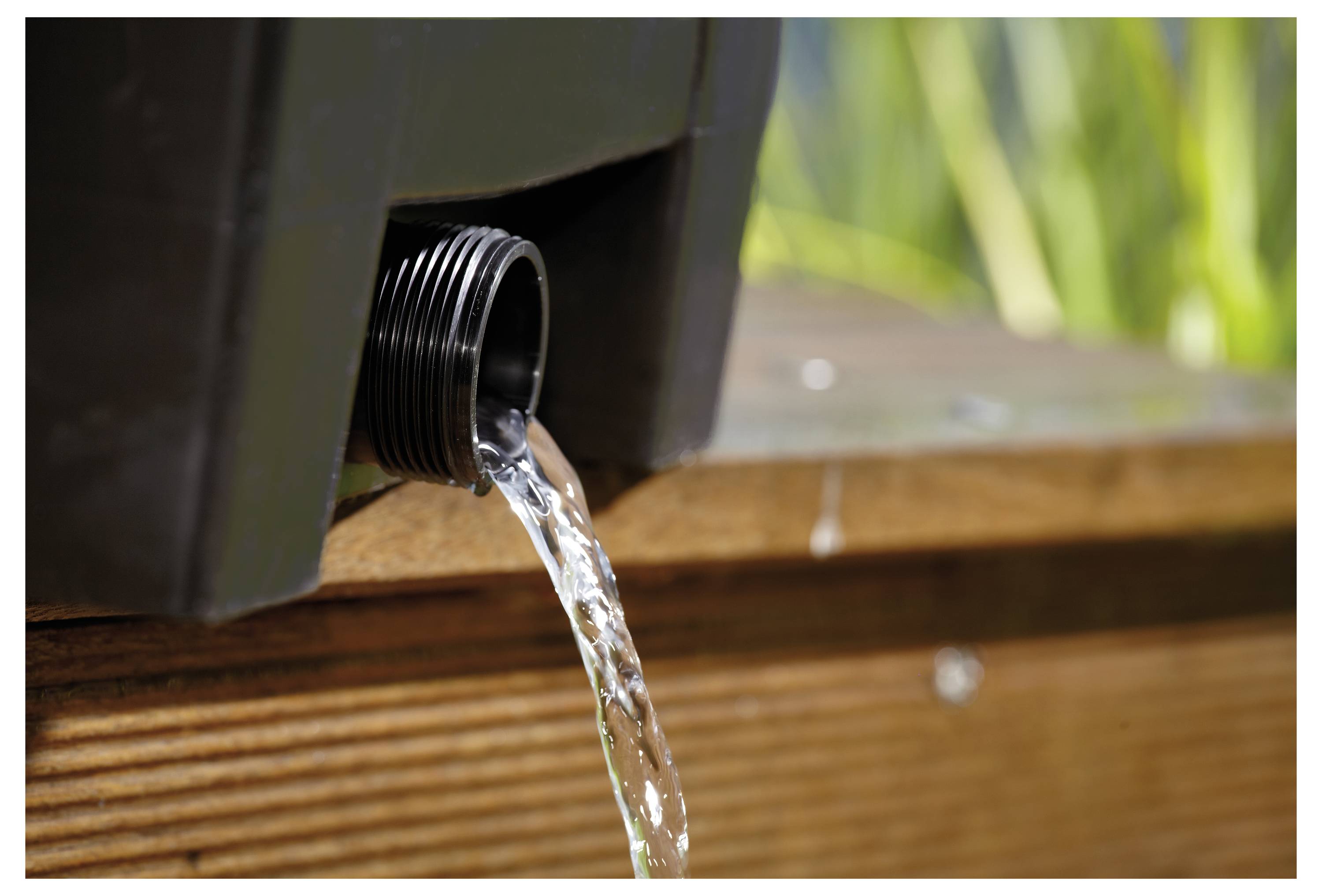 Wasser fließt aus einer Öffnung am Boden eines dunklen Behälters und zeigt eine kontrollierte Freisetzung auf einer Holzoberfläche im Freien.