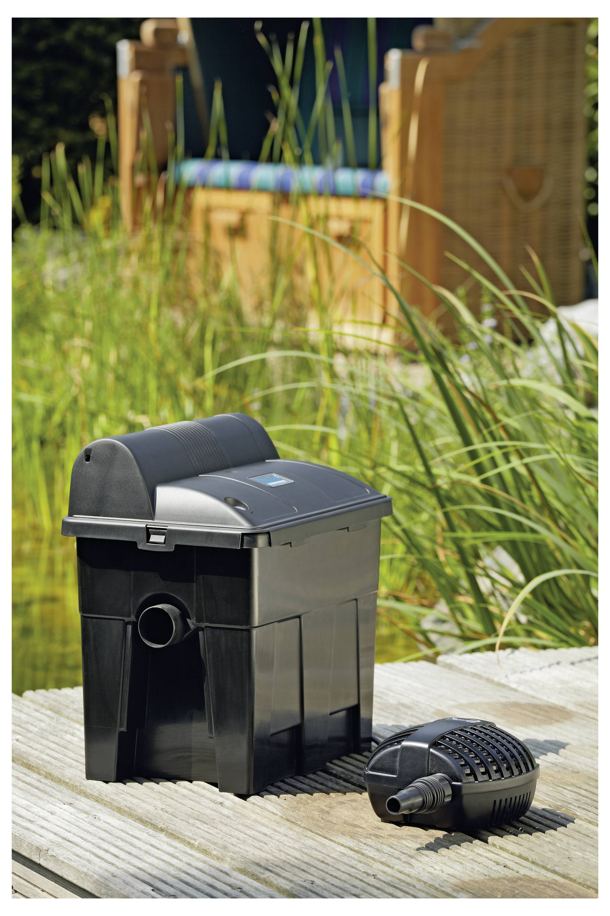 Ein Teichfilter und eine Pumpe stehen auf einer Holzterrasse in der Nähe eines Gartenteiches mit hohem Gras und einer Bank im Hintergrund, die zur Wasserfilterung verwendet werden.