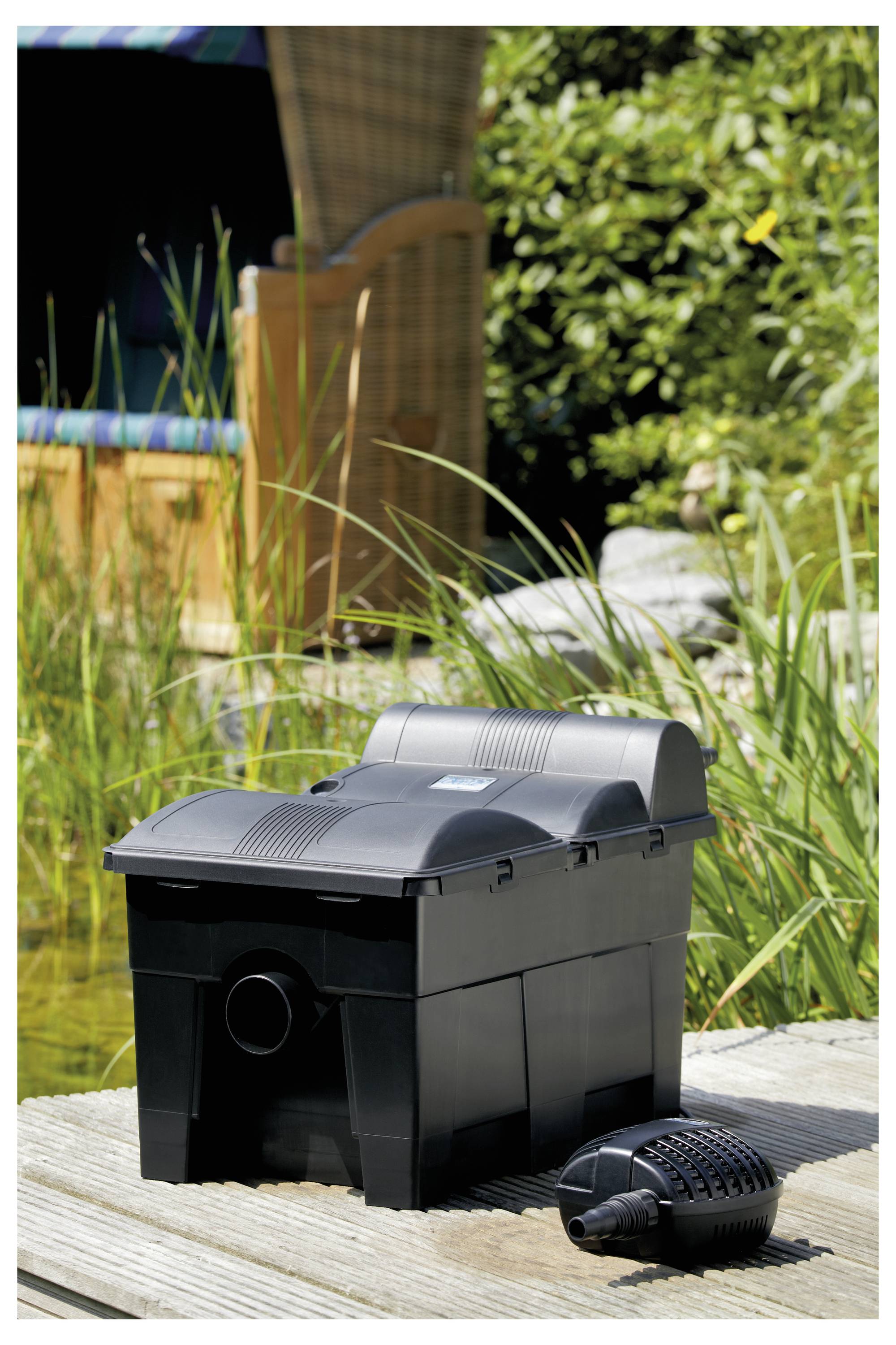 Teichreinigungseinheit mit Pumpe auf einem Holzdeck neben einem Teich, umgeben von Grünpflanzen und einem Korbstuhl im Hintergrund.