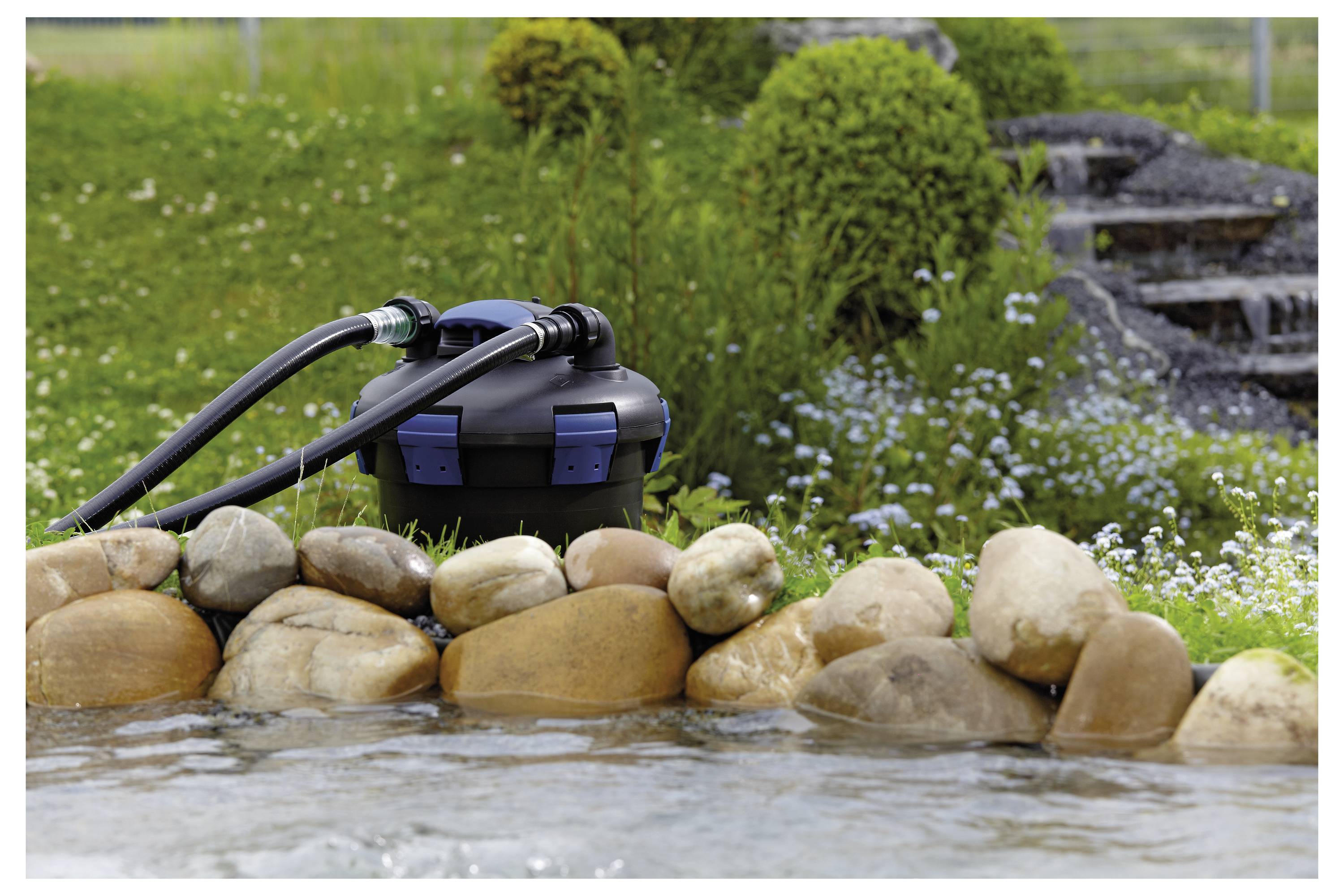 Teichfilter mit Schläuchen neben einem steingefassten Teich, umgeben von Gras und Blumen, mit einem kleinen Wasserfall im Hintergrund.