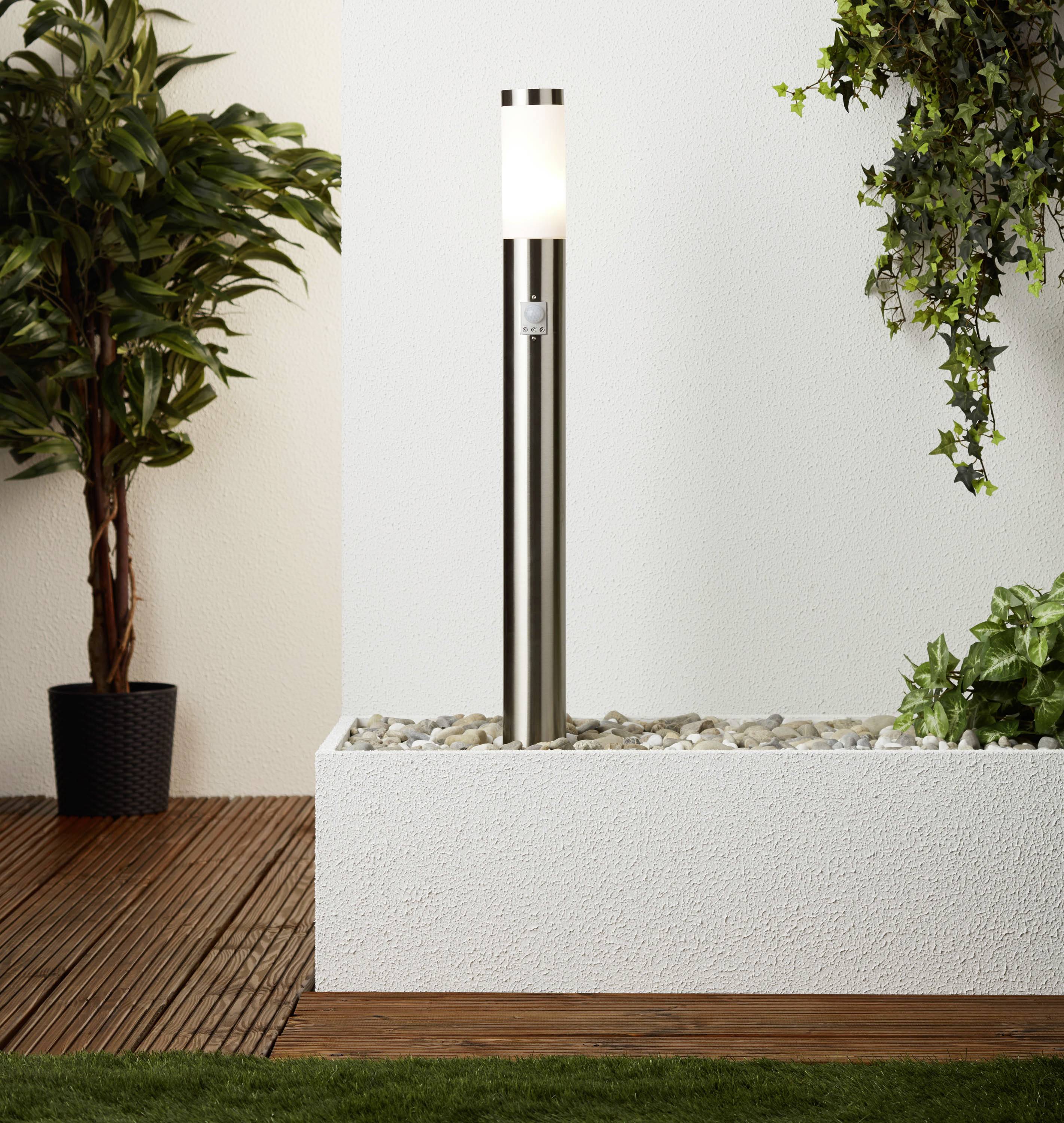 Eine moderne Außenleuchte aus Edelstahl steht auf einem Kiesbett, umgeben von Pflanzen. Sie beleuchtet eine Terrasse mit Holz- und Grasdetails.