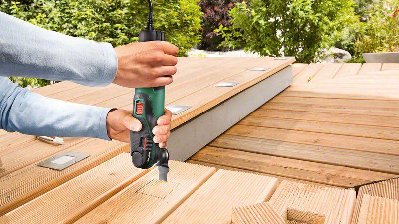 Eine Person benutzt ein elektrisches Multitool, um einen rechteckigen Ausschnitt in einer Holzterrasse zu schneiden.