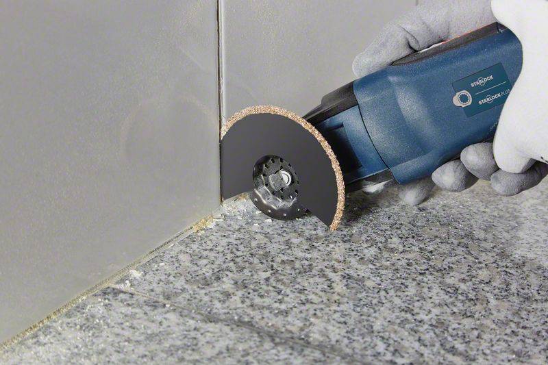 Hand mit Handschuh hält ein elektrisches Multifunktionswerkzeug mit rotierender Klinge, schneidet Fliesen an der Wand entlang.