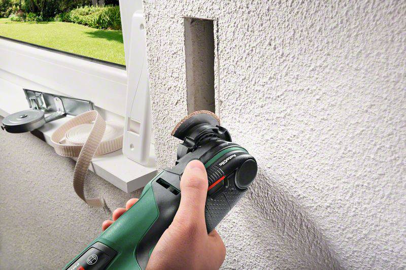 Eine Hand hält ein Multitool, das ein rechteckiges Loch in eine Wand schneidet, vermutlich für den Einbau eines Schalters oder Steckdose.