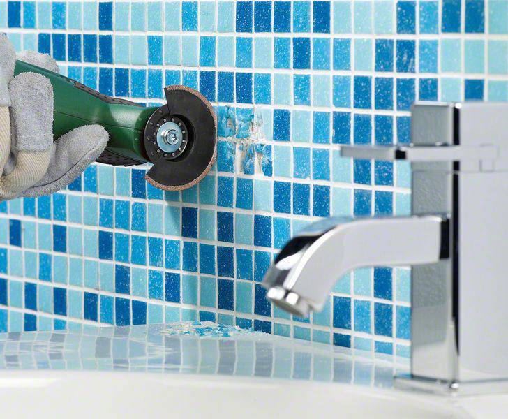 Eine Person verwendet ein Elektrowerkzeug, um blaue Mosaikfliesen an einer Wand zu schneiden. Im Vordergrund ist ein Wasserhahn sichtbar.