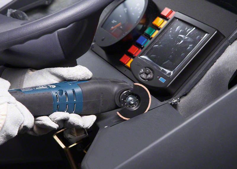 Eine mit Handschuhen geschützte Hand benutzt ein Elektrowerkzeug, um ein Armaturenbrett mit einem Bildschirm und bunten Tasten zu bearbeiten.