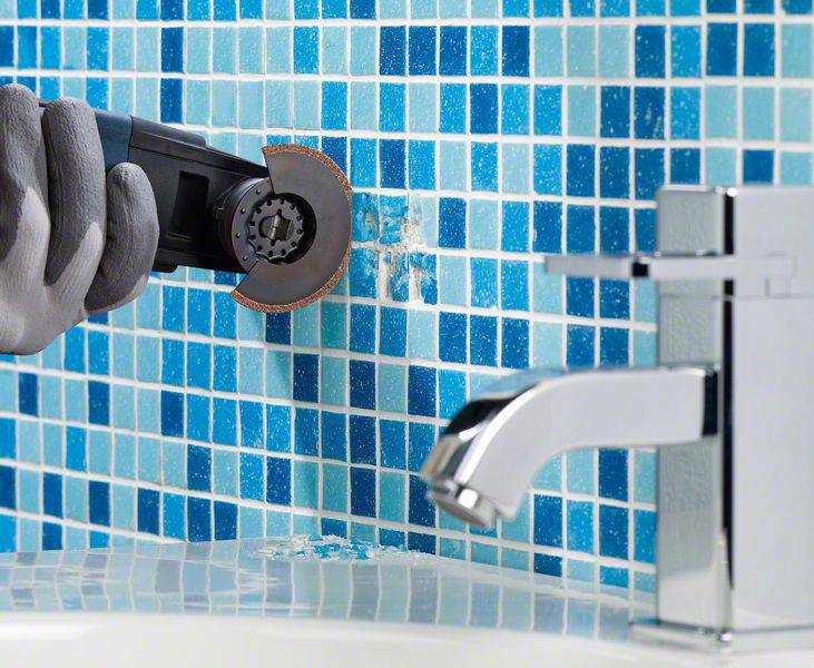 Eine behandschuhte Hand hält ein Elektrowerkzeug und entfernt beschädigte Fliesen von einer blauen Mosaikwand neben einem Wasserhahn.