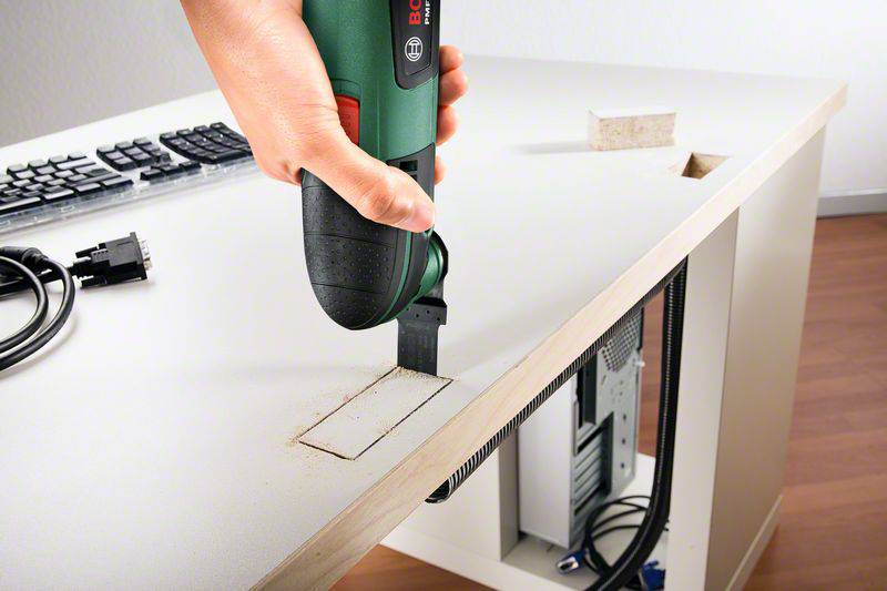 Eine Hand hält ein Elektrowerkzeug, um ein rechteckiges Loch in eine Tischplatte zu schneiden. Kabel und ein Computer sind sichtbar.