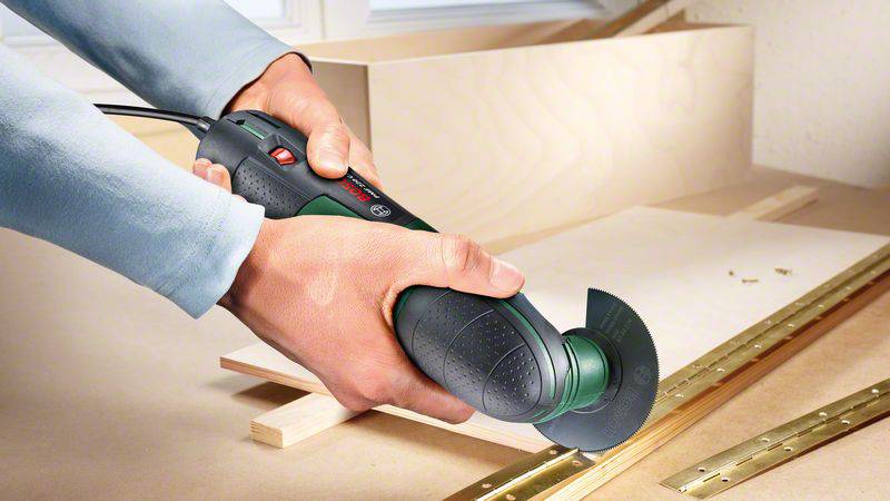 Eine Person schneidet präzise ein Holzstück mit einem elektrischen Multifunktionstool. Schrauben und Holzleisten liegen daneben.
