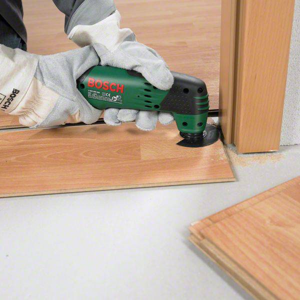 Eine Person schneidet mit einem grünen Bosch-Multifunktionswerkzeug einen Holzboden an einer Türschwelle. Hände sind in Arbeitshandschuhen.