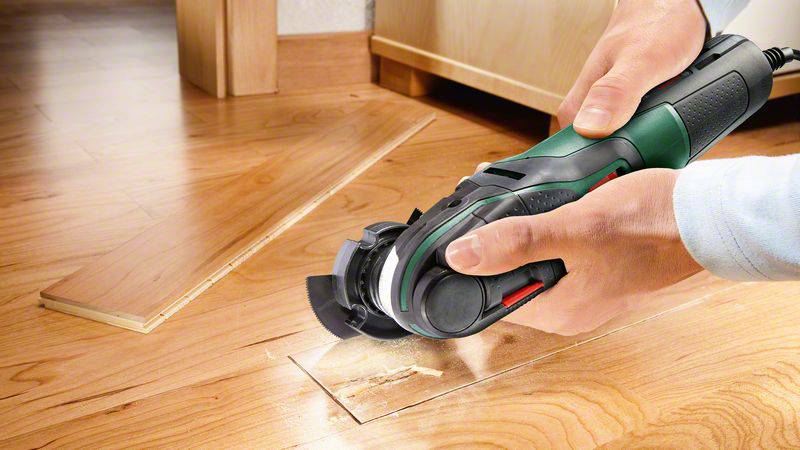 Eine Person schneidet mit einem elektrischen Werkzeug eine Holzplatte. Auf dem Boden liegen bereits geschnittene Holzstücke.