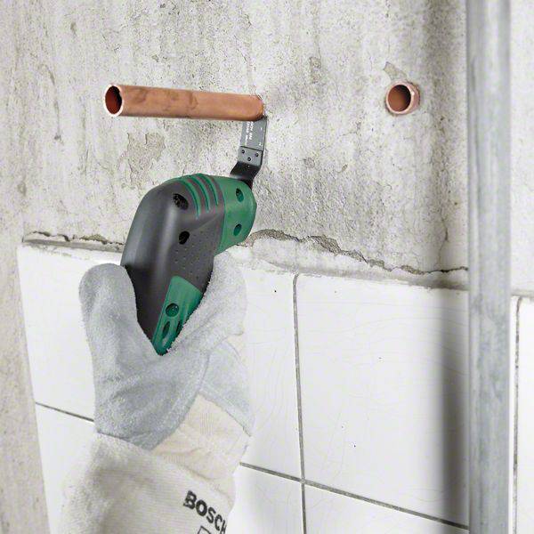 Eine Person bohrt in eine Wand mit einem grünen Akkuschrauber. Zwei Kupferrohrenden ragen aus der Wand.