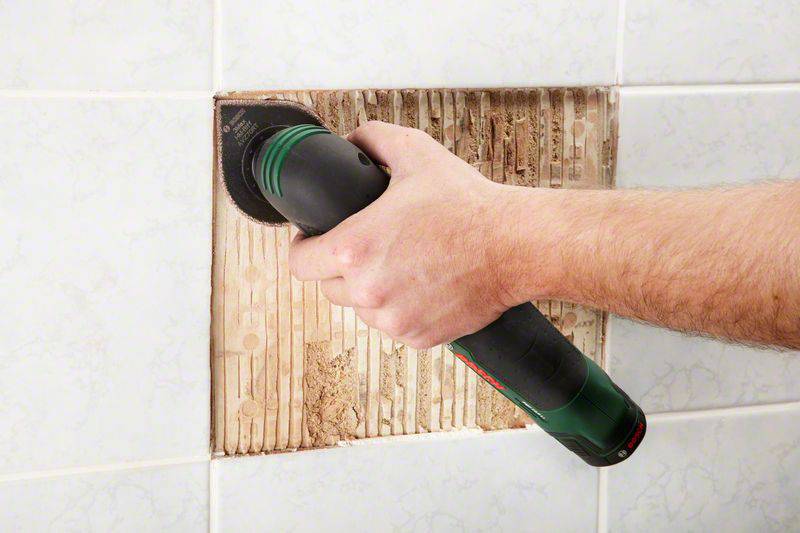 Eine Person entfernt mit einem Elektrowerkzeug Kacheln von einer Wand. Teile des darunterliegenden Materials sind sichtbar.