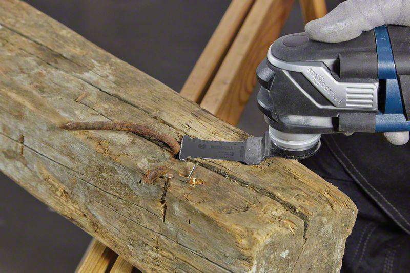 Ein oszillierendes Werkzeug entfernt vorsichtig einen alten, rostigen Nagel aus einem groben Holzbalken.