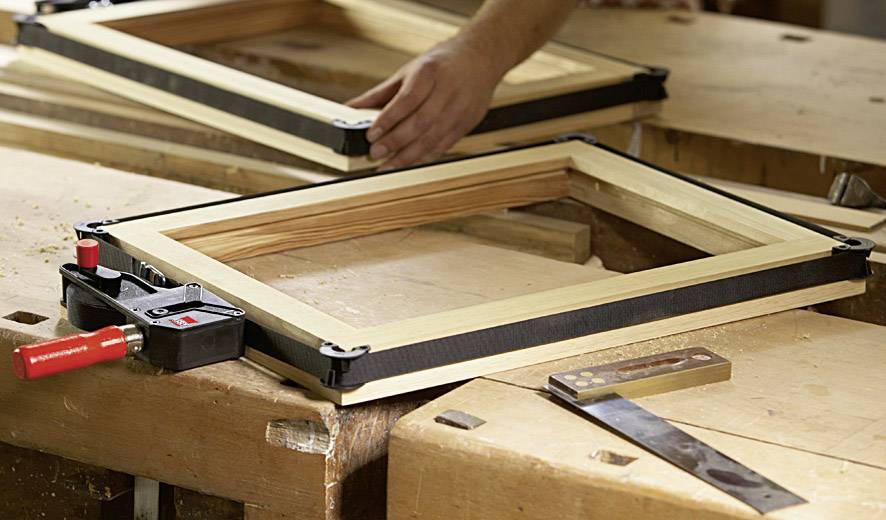 Ein Handwerker baut einen Holzrahmen auf einer Werkbank zusammen, umgeben von Werkzeug wie einem Schraubendreher und einem Winkelmesser.
