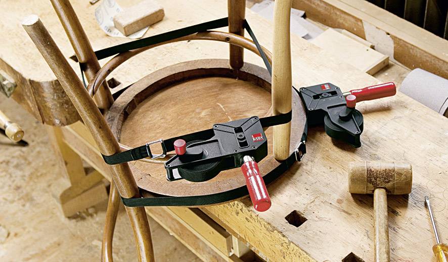 Eine Werkbank mit einem Holzstuhlrahmen in Bearbeitung. Zwei Spanngurte befestigen die runden Holzstücke. Ein Holzhammer liegt daneben.