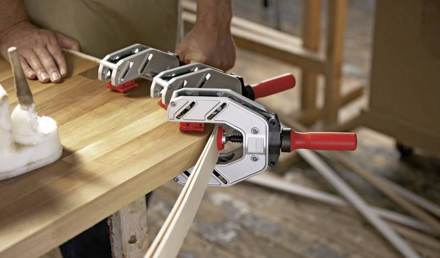 Eine Person benutzt drei Klemmen, um einen gebogenen Holzstreifen an einer Tischplatte zu befestigen. Die Szene zeigt eine handwerkliche Tätigkeit.