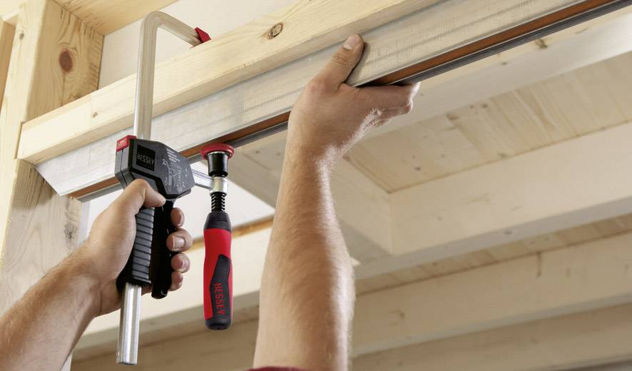 Zwei Hände verwenden eine Schraubzwinge, um zwei Holzbalken zusammenzufügen. Die Szene zeigt präzises Arbeiten mit Holz.