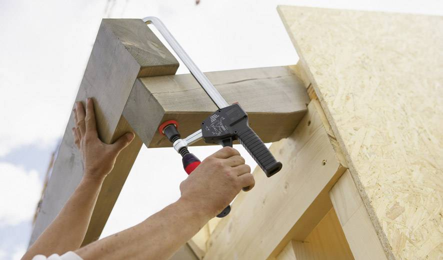 Zwei Hände halten Holzbalken mit einer großen Zwinge zusammen. Eine Konstruktion aus Holz wird errichtet, möglicherweise ein Dachstuhl.