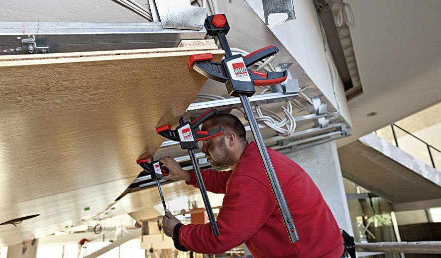 Ein Handwerker in roter Jacke befestigt mit Schraubzwingen eine Holzplatte an einer Decke, um sie zu montieren.