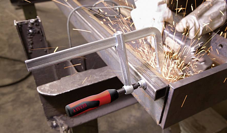Eine Schraubzwinge hält Metallteile fest, während ein Arbeiter mit einem Winkelschleifer Funken sprüht. Bearbeitung in einer Werkstatt.
