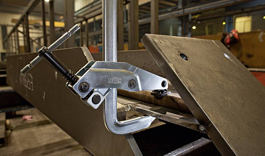 Industriehalle mit Metallarbeiten: Ein C-Klemme fixiert ein Metallstück an einem Stahlträger. Maschinen und Werkzeuge im Hintergrund.