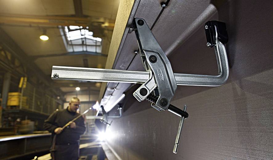 Ein Monteur arbeitet in einer Werkstattumgebung. Im Vordergrund ist eine große Metallklemme sichtbar, die an einem Werkstück befestigt ist.