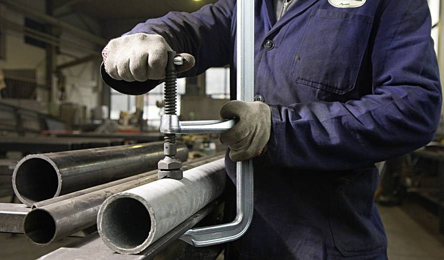 Eine Person in Arbeitskleidung fixiert Metallrohre in einer Werkstatt mit einer großen Schraubzwinge.