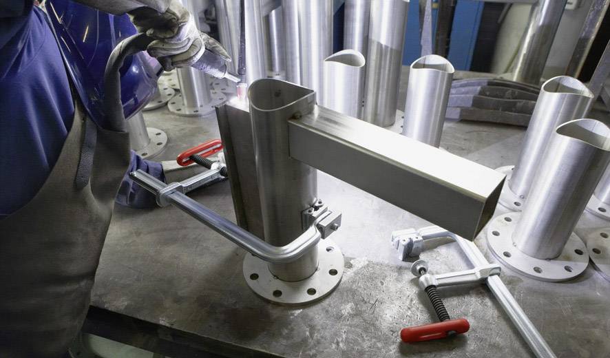 Eine Person schweißt ein Stahlrohr in einer Werkstatt. Metallteile und Zwingen sind auf dem Tisch verteilt.