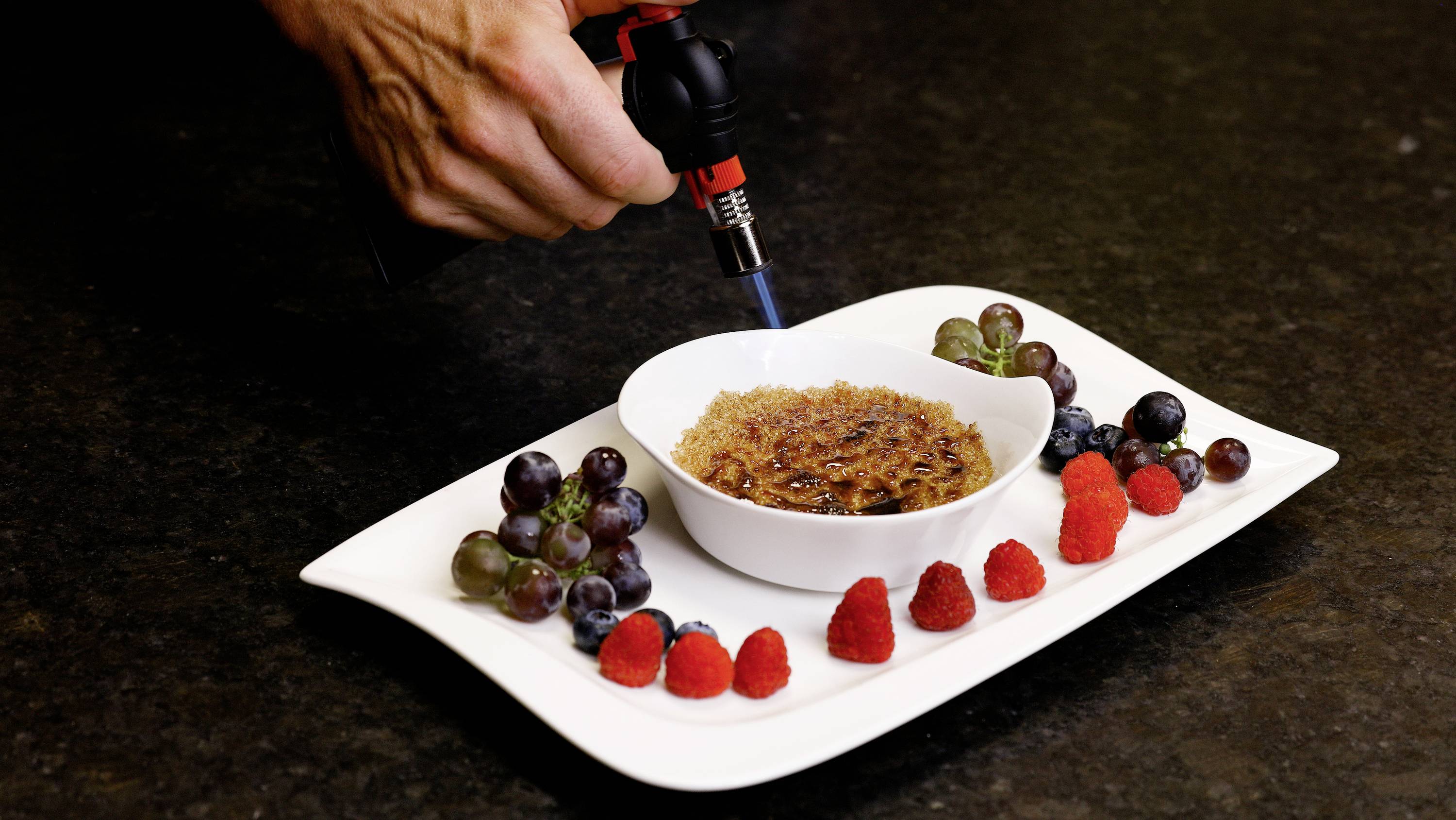 Eine Hand karamellisiert die Zuckerkruste auf einer Crème brûlée mit einem Bunsenbrenner. Der Teller ist mit frischen Trauben und Himbeeren garniert.